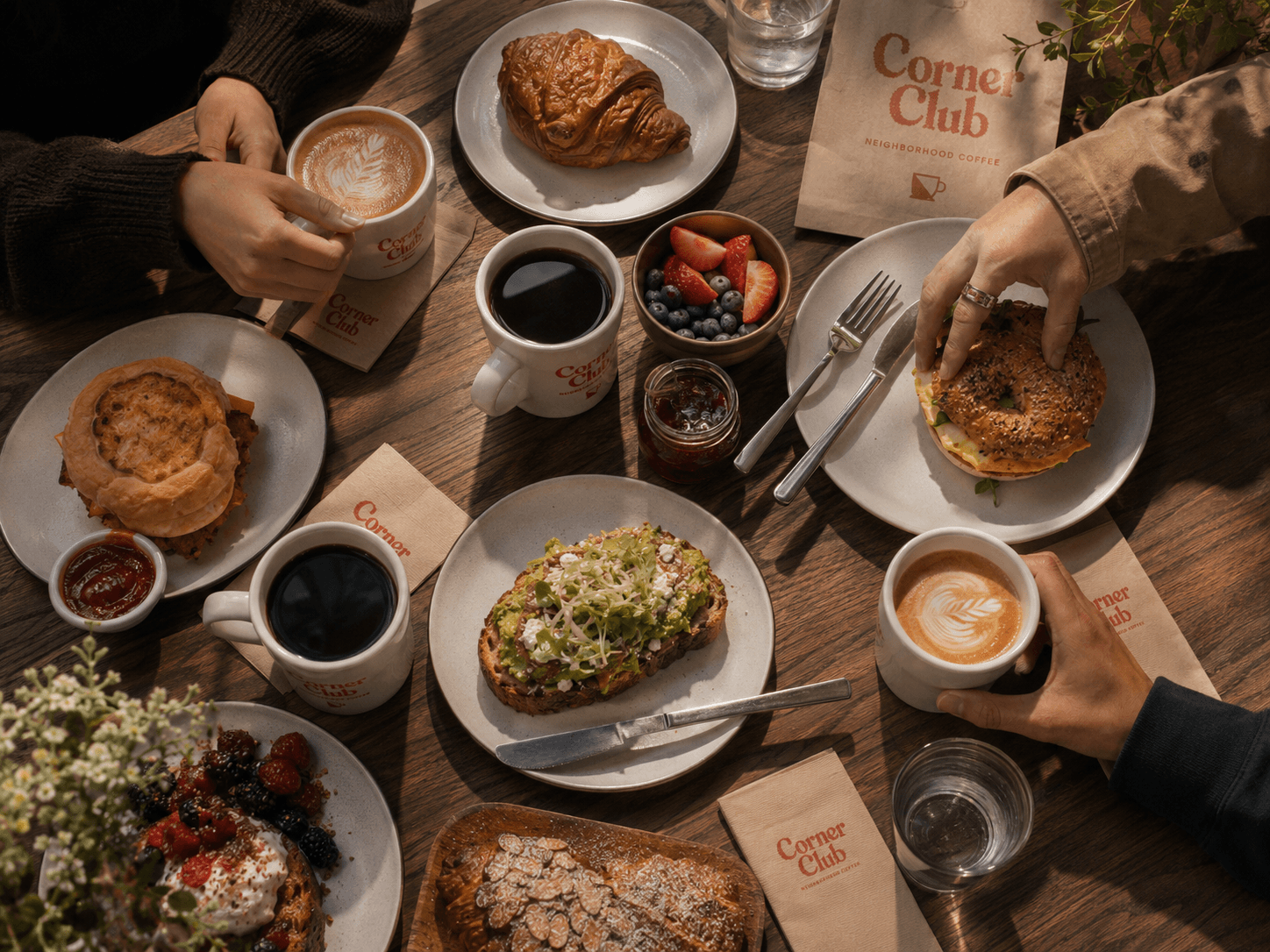 Overhead spread of Corner Club coffee, avocado toast, croissants and berries on a wooden table